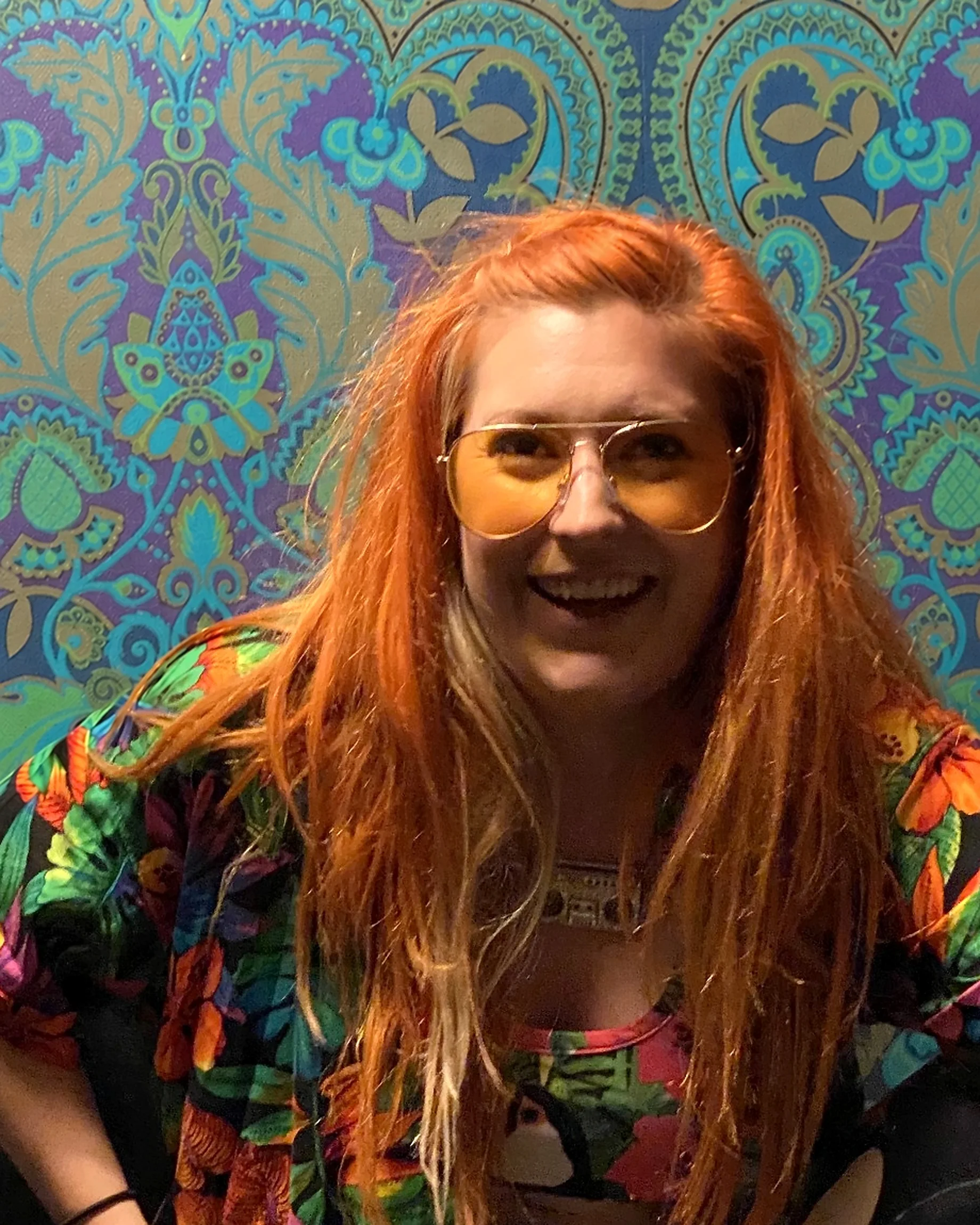 Vicki smiling in front of a colourful patterned wall, wearing a bright floral outfit and amber glasses.